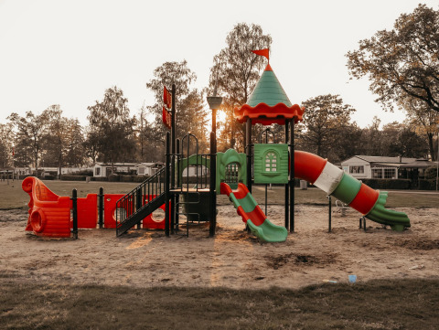 Parque infantil colorido con toboganes y arena, junto a árboles y cabañas glamping al atardecer.
