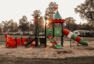 Parque infantil colorido con toboganes y arena, junto a árboles y cabañas glamping al atardecer.