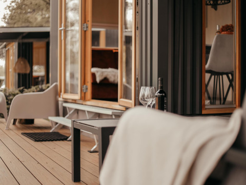 Acogedora terraza exterior de una cabaña moderna con ventanas abiertas, vino y cómodas sillas.