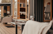 Acogedora terraza exterior de una cabaña moderna con ventanas abiertas, vino y cómodas sillas.