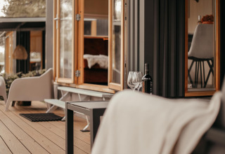 Acogedora terraza exterior de una cabaña moderna con ventanas abiertas, vino y cómodas sillas.