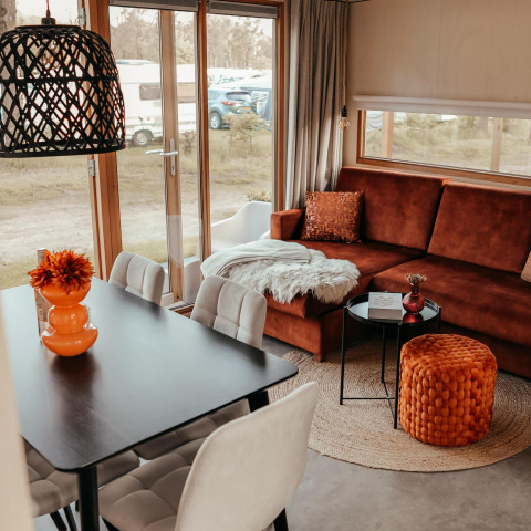 Interior moderno de cabaña con sofá marrón, puf naranja, mesa de comedor y grandes ventanales a la zona de camping.