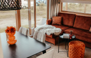 Interior moderno de cabaña con sofá marrón, puf naranja, mesa de comedor y grandes ventanales a la zona de camping.