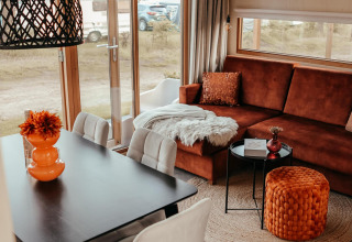 Interior moderno de cabaña con sofá marrón, puf naranja, mesa de comedor y grandes ventanales a la zona de camping.