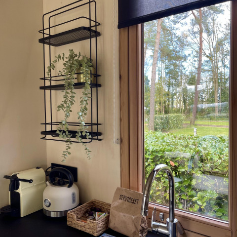 Rincón de cocina en una cabaña con cafetera, tetera, estante con planta y vista al jardín arbolado.