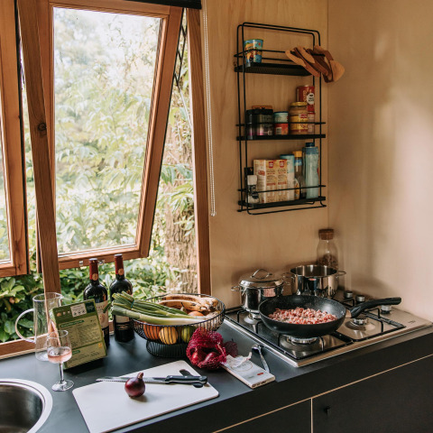 Cuisine chaleureuse de cabine avec fenêtre ouverte, légumes frais, vin et cuisson en cours sur la cuisinière.