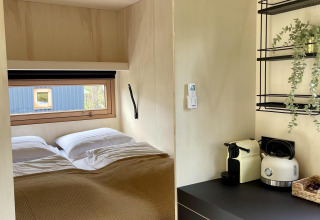 Acogedora cabaña con cama doble, ventana, pequeña cocina con cafetera, hervidor y planta decorativa.