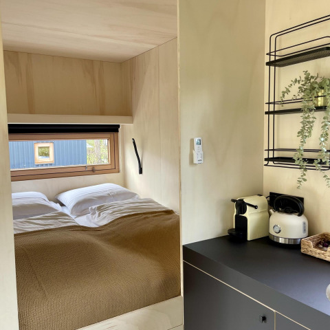 Acogedora cabaña con cama doble, ventana, pequeña cocina con cafetera, hervidor y planta decorativa.