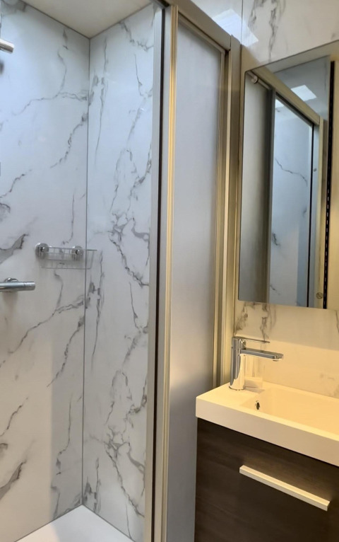 Baño moderno de cabina con paredes tipo mármol, ducha, lavabo y espejo, iluminado por luz natural.