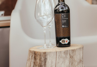 Botella de vino y dos copas sobre un tronco de árbol frente a un sillón en una cabaña acogedora.