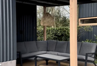 Terraza exterior de una cabaña con sofá gris en L, mesa, lámpara colgante y vistas al jardín arbolado.
