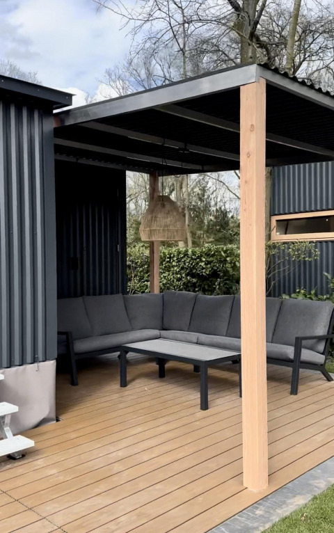 Terraza exterior de una cabaña con sofá gris en L, mesa, lámpara colgante y vistas al jardín arbolado.