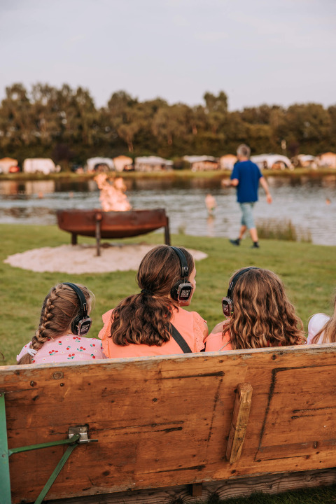 Three girls wearing headphones sit on a bench by a lake at Recreatiepark De Achterste Hoef.