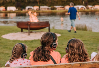 Tre piger med hovedtelefoner sidder på en bænk ved en sø i Recreatiepark De Achterste Hoef.