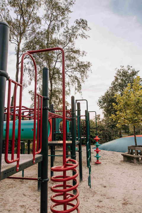 Spielplatz mit Klettergerüst und Rutsche im Sand, umgeben von Bäumen in De Achterste Hoef, Niederlande.
