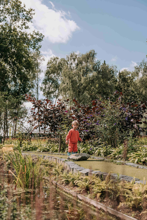 Een kind in oranje kledij bij een vijver met planten in Recreatiepark De Achterste Hoef, Noord-Brabant.