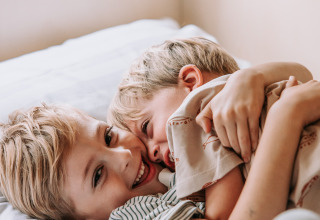 Due bambini felici si abbracciano e sorridono insieme su un letto a Recreatiepark De Achterste Hoef.