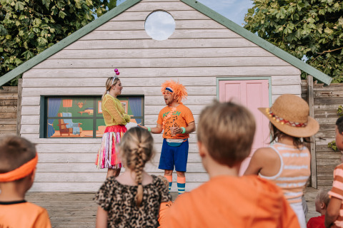 Children watch a colorful outdoor play in front of a wooden house at De Achterste Hoef holiday park.