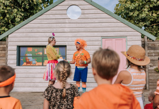 Børn ser på et farverigt udendørs teaterstykke foran et træhus i ferieparken De Achterste Hoef.