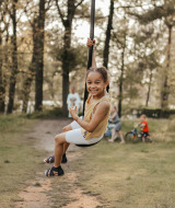 Una niña sonriente se desliza en una tirolesa en Recreatiepark De Achterste Hoef, rodeada de árboles.