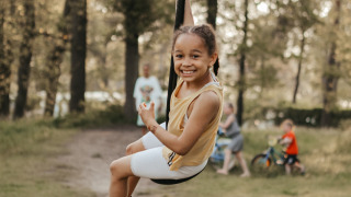 Una niña sonriente se desliza en una tirolesa en Recreatiepark De Achterste Hoef, rodeada de árboles.