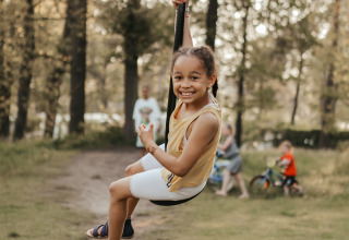 Et smilende barn gynger på en zipline i Recreatiepark De Achterste Hoef, omgivet af skov og familier.