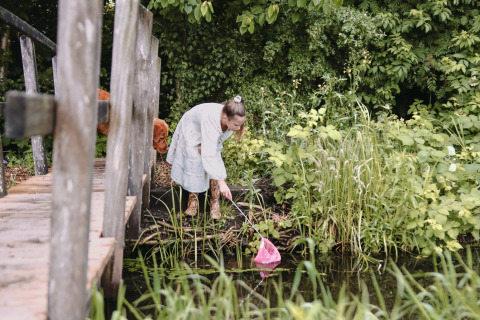 Vrouw in witte jurk schept met netje aan een vijver, groene natuur, Feather Down Botterheerd, Groningen.