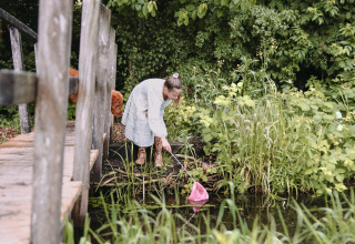 Vrouw in witte jurk schept met netje aan een vijver, groene natuur, Feather Down Botterheerd, Groningen.