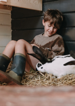 Niño con botas de goma sentado en paja junto a conejos en Feather Down Botterheerd, Groningen, Países Bajos.