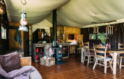 Interieur van een sfeervolle safaritent met houtkachel, eettafel, keukengedeelte en rustieke meubels.