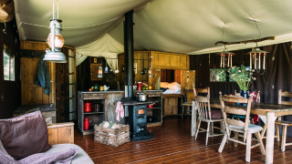 Interieur van een sfeervolle safaritent met houtkachel, eettafel, keukengedeelte en rustieke meubels.