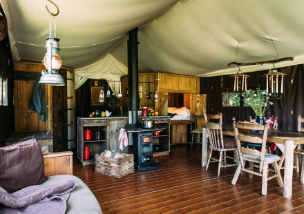 Interieur van een sfeervolle safaritent met houtkachel, eettafel, keukengedeelte en rustieke meubels.