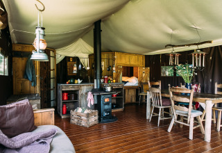 Interior of a cozy safari tent featuring a wood stove, dining table, kitchen area, and rustic furnishings.