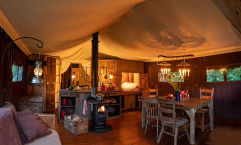 Interno di tenda safari con illuminazione calda, stufa a legna, cucina rustica e tavolo da pranzo in legno.