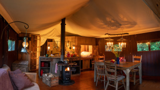 Safari tent interior with warm lighting, wood-burning stove, rustic kitchen, dining table and wooden furniture.