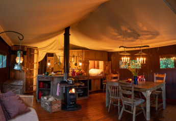 Interior de tienda safari con iluminación cálida, estufa de leña, cocina rústica y mesa de comedor de madera.
