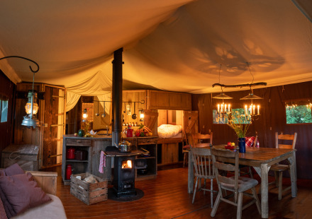 Safari tent interieur met warme verlichting, houtkachel, rustieke keuken en houten eettafel en stoelen.