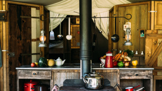 Rustic safari tent kitchen with wooden cabinets, fresh vegetables, hanging utensils, and wood stove.