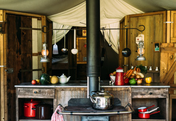 Cucina rustica in tenda safari con mobili in legno, verdure fresche e utensili sospesi a vista.