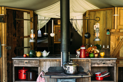 Cucina rustica in tenda safari con mobili in legno, verdure fresche e utensili sospesi a vista.