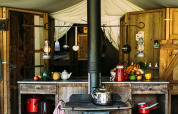 Rustic safari tent kitchen with wooden cabinets, fresh vegetables, hanging utensils, and wood stove.