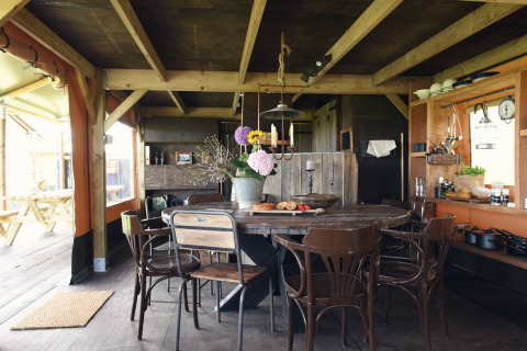 Interno di una sistemazione glamping con tavolo rotondo in legno, sedie diverse e cucina rustica a vista.
