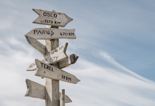 Panneau en bois dans un glamping indiquant les distances vers Oslo, Paris, New York et Texel, ciel bleu.