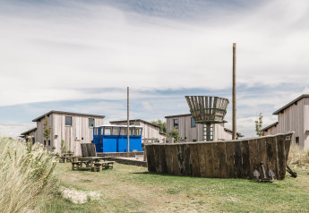 Alojamiento glamping con cabañas de madera, autobús azul y barco de juego de madera al aire libre, rodeado de naturaleza.