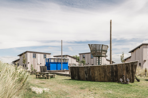 Glampingverblijf met houten chalets, blauwe bus en speels houten schip als speeltoestel in het groen.