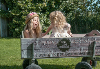 Dos niñas ríen y juegan en un carro de madera sobre el césped en el glamping Duynpark het Zwanenwater.