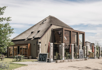 Photo of a modern building with thatched roof and glass front at a glamping site with outdoor seating.