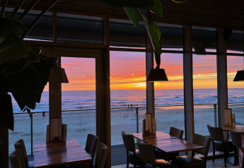 Restaurante de glamping con mesas de madera y vista al mar a través de ventanales durante el atardecer.