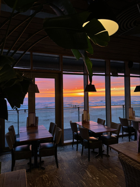 Glamping dining area with wooden tables and chairs facing large windows overlooking a sunset beach view.