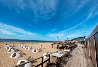 Glamping en la playa con tiendas modernas, huéspedes relajados y restaurante con terraza frente al mar soleado.